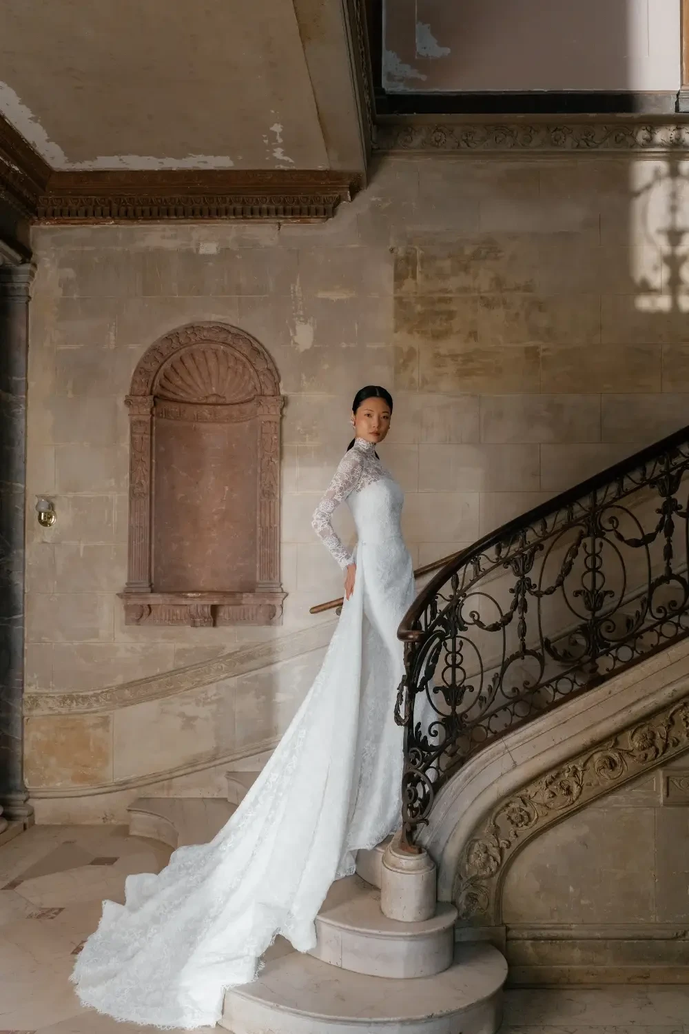 A woman in a white dress standing on a staircase with ornate railings in a vintage-style setting.