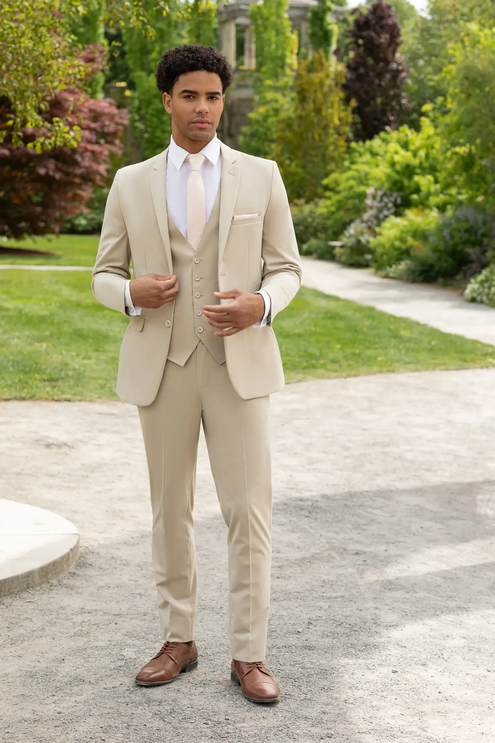 A man wearing a beige suit with a vest and light pink tie, standing in a landscaped garden setting.