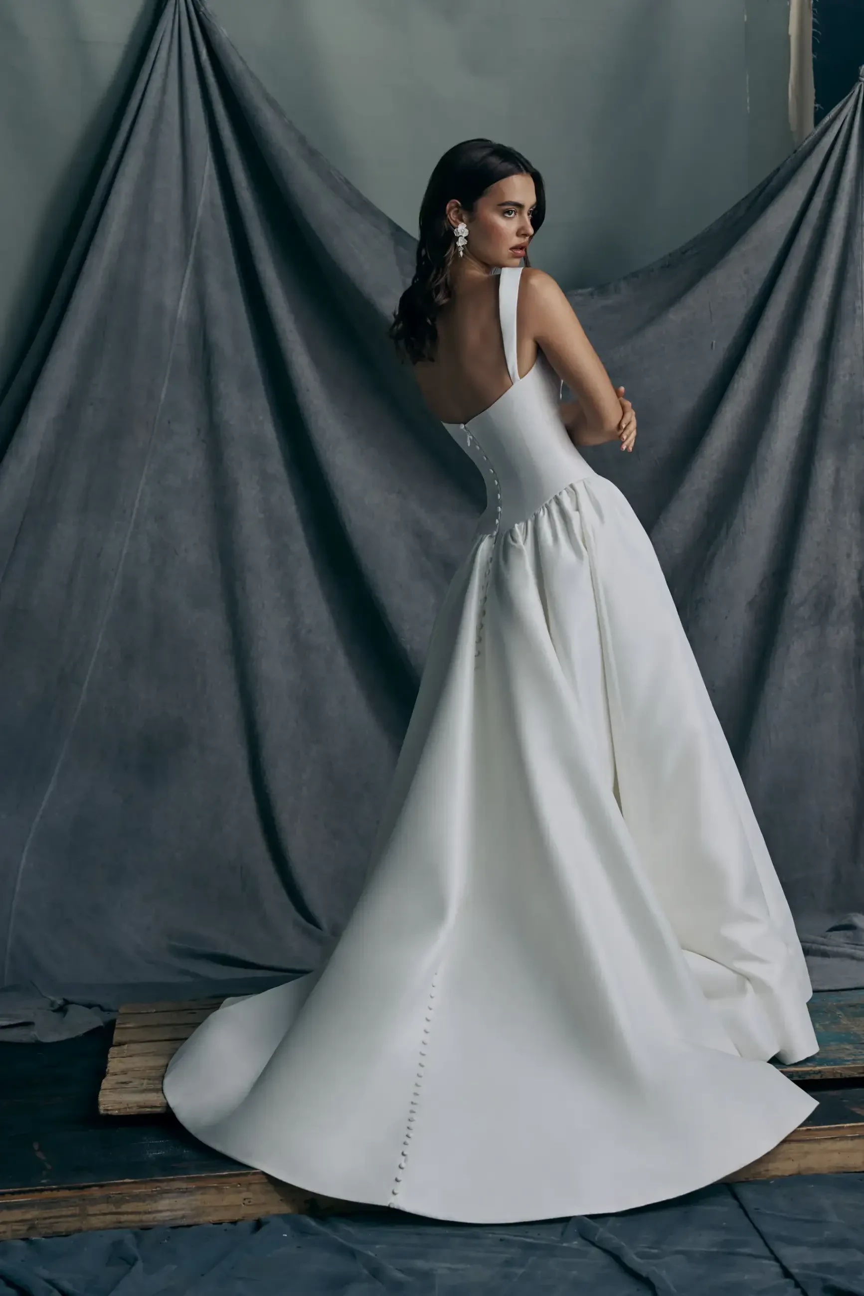 A model is wearing a white wedding gown with a fitted bodice and flowing train, standing against a textured backdrop.