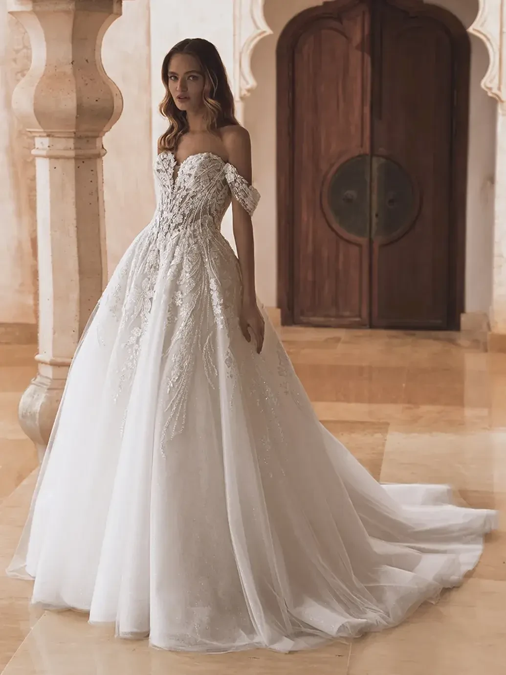 A woman in an elegant off-shoulder wedding dress with intricate detailing, standing gracefully in a beautifully designed interior space.