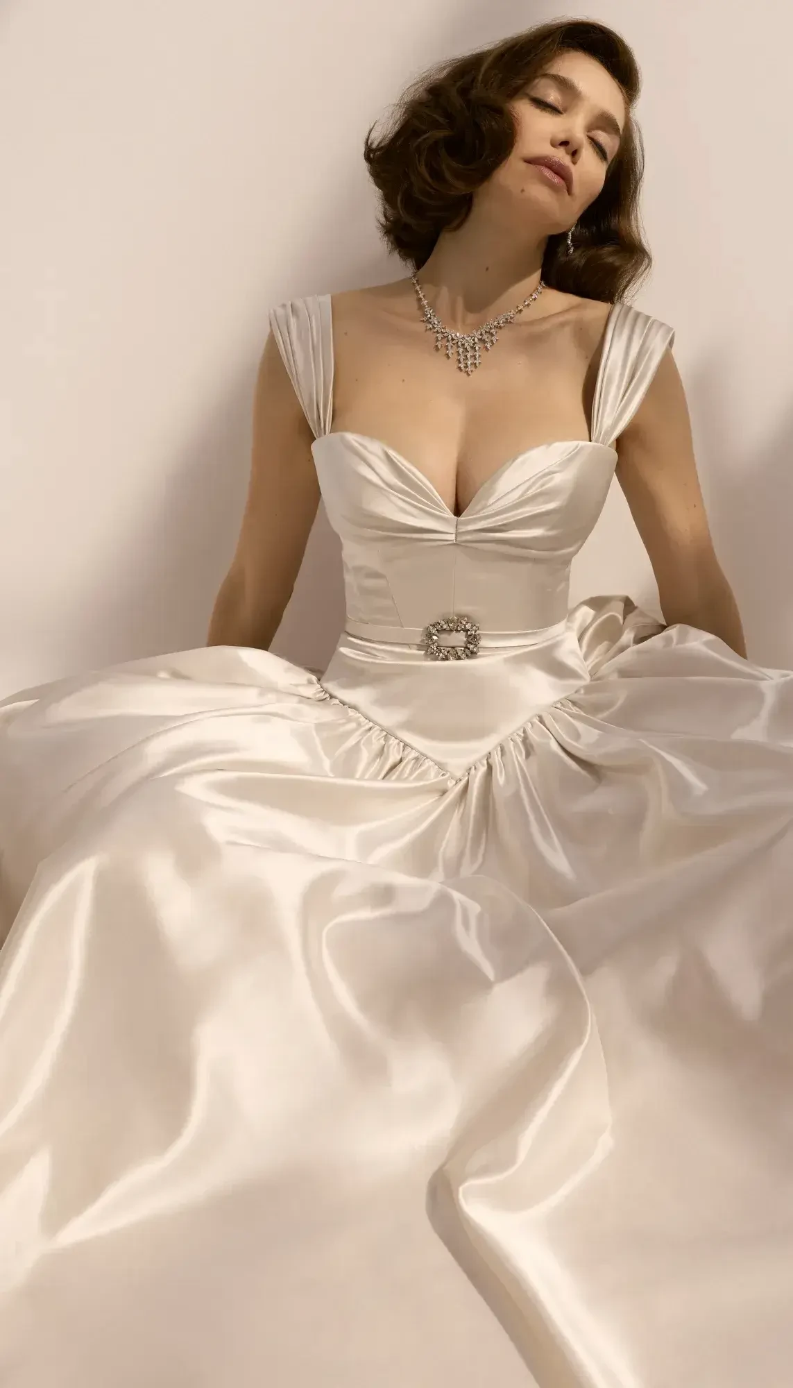 A woman in an elegant satin gown with a plunging neckline, sitting gracefully against a neutral background, wearing a delicate necklace.